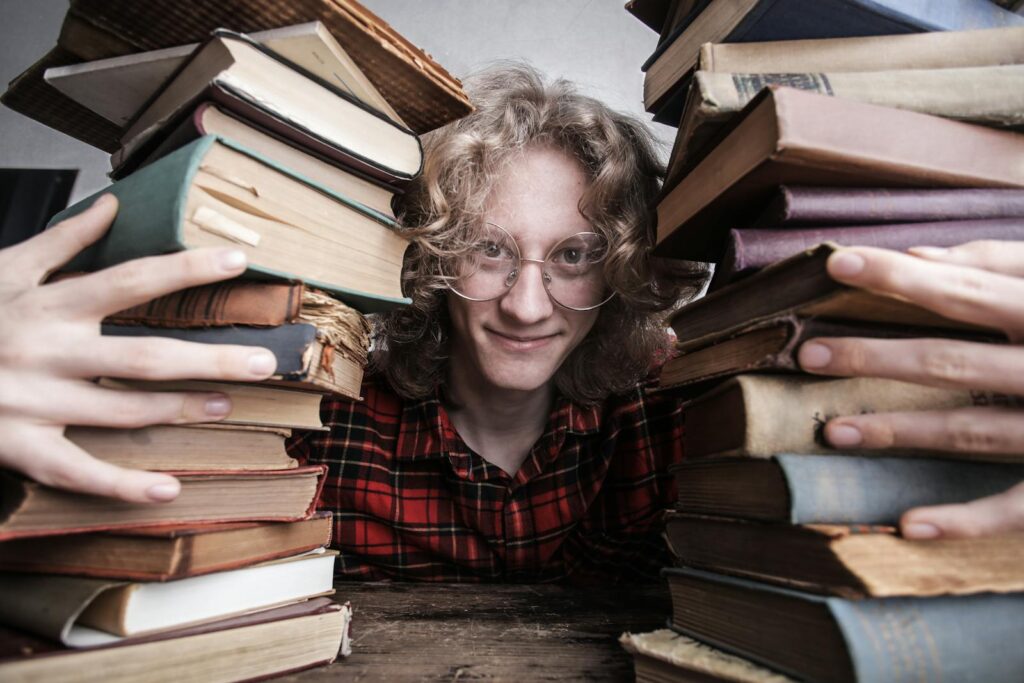 A young man in glasses surrounded by stacks of vintage books indoors, embodying knowledge and learning.