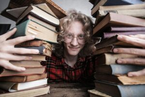 A young man in glasses surrounded by stacks of vintage books indoors, embodying knowledge and learning.